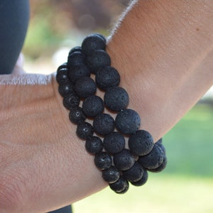 May include: Three black lava rock beaded bracelets stacked on a person's wrist.