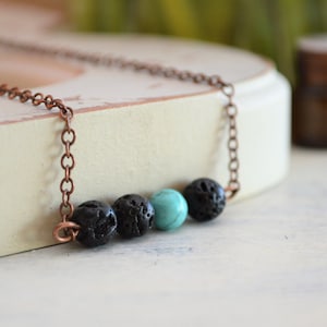 Puede incluir: Un collar de cadena de cobre con cuentas de roca volcánica negra y una cuenta turquesa. El collar está sobre una superficie blanca.