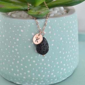 May include: A rose gold necklace with a black lava stone pendant and a circular charm engraved with the letter "K". The necklace is displayed in front of a light blue pot with white polka dots, holding a green succulent.