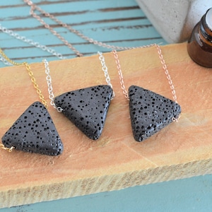 May include: Four lava stone triangle pendants on silver, gold, and rose gold chains. The pendants are black and have a rough, textured surface. A small brown glass bottle with a black cap is in the background.