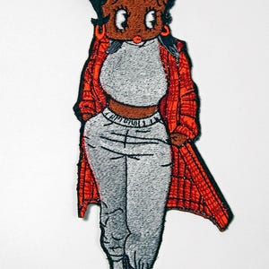 Peut inclure: Écusson brodé représentant un personnage de dessin animé vêtu d'un crop top gris, d'un pantalon de survêtement assorti et d'une veste à carreaux rouge. Le personnage a la peau foncée, des cheveux noirs et des chaussures rouges. Sur fond blanc.