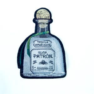 May include: Embroidered patch in the shape of a tequila bottle. The patch features the words "Silver Patron" and "Tequila 100% de Agave". The bottle is light blue with a green ribbon and a cork top.