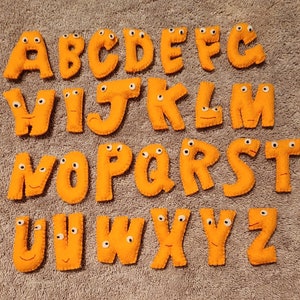 Puede incluir: Letras del alfabeto de fieltro naranja con ojos saltones. Cada letra está cosida individualmente y tiene una expresión caprichosa y juguetona.