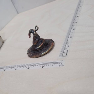 Peut inclure: Un petit chapeau de sorcier en m&eacute;tal brun fonc&eacute; avec un sommet pointu et un bord recourb&eacute;. Le chapeau a une boucle sur le dessus pour l'accrocher.