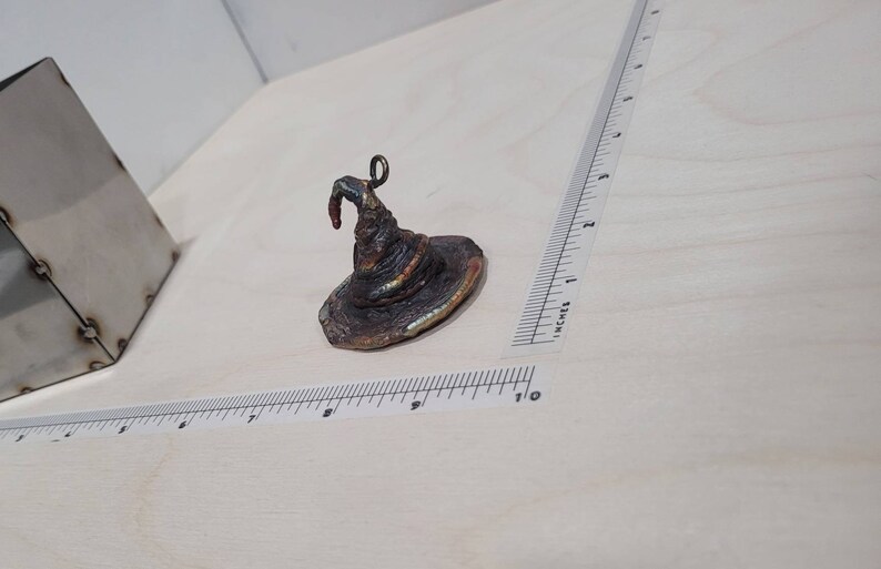 Peut inclure: Un petit chapeau de sorcier en m&eacute;tal brun fonc&eacute; avec un sommet pointu et un bord recourb&eacute;. Le chapeau a une boucle sur le dessus pour l'accrocher.
