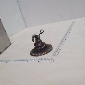 Peut inclure: Un petit chapeau de sorcier en m&eacute;tal brun fonc&eacute; avec un bord recourb&eacute; et une petite boucle en m&eacute;tal sur le dessus. Le chapeau est pos&eacute; sur une surface blanche.