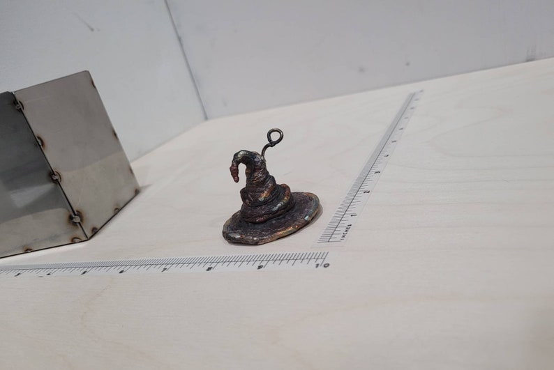 Peut inclure: Un petit chapeau de sorcier en m&eacute;tal brun fonc&eacute; avec un bord recourb&eacute; et une petite boucle en m&eacute;tal sur le dessus. Le chapeau est pos&eacute; sur une surface blanche.