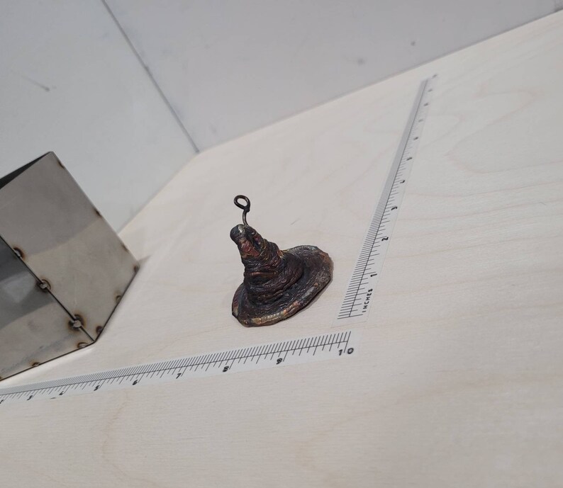 Peut inclure: Un petit chapeau de sorcier en c&eacute;ramique brun avec une boucle de fil sur le dessus. Le chapeau est pos&eacute; sur une surface blanche.