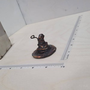 Peut inclure: Une petite sculpture artisanale en cuivre de couleur cuivre du Choixpeau magique de la s&eacute;rie Harry Potter. Le chapeau est pos&eacute; sur une surface blanche.