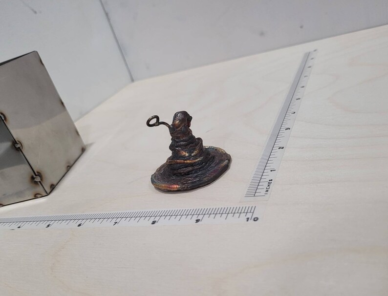 Peut inclure: Une petite sculpture artisanale en cuivre de couleur cuivre du Choixpeau magique de la s&eacute;rie Harry Potter. Le chapeau est pos&eacute; sur une surface blanche.