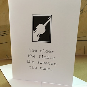 Puede incluir: Tarjeta de felicitación blanca con una silueta de violín en blanco y negro dentro de un rectángulo negro. El texto dice: "Cuanto más viejo el violín, más dulce la melodía". La tarjeta está dentro de un sobre blanco.