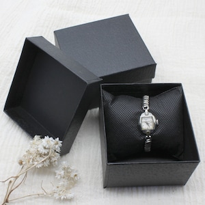 Vintage silver wristwatch in the basic box option. The watch is holding a diamond design black fabric pillow inside a black hard cardboard, leather textured box, with separate cover. They are sitting on white linen with dried white flowers in front