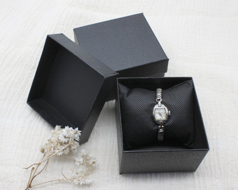 Vintage silver wristwatch in the basic box option. The watch is holding a diamond design black fabric pillow inside a black hard cardboard, leather textured box, with separate cover. They are sitting on white linen with dried white flowers in front