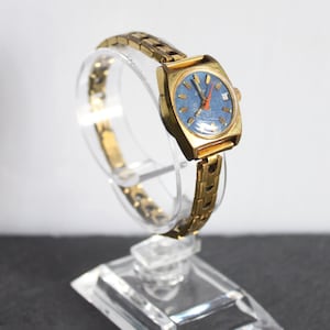 Side view of a functional vintage 1970 gold Latendresse womens mechanical wind up wristwatch. This Stylish watch has a blue dial with golden markers and a contrasting orange hand. It is on a clear bracelet display on white and grey background