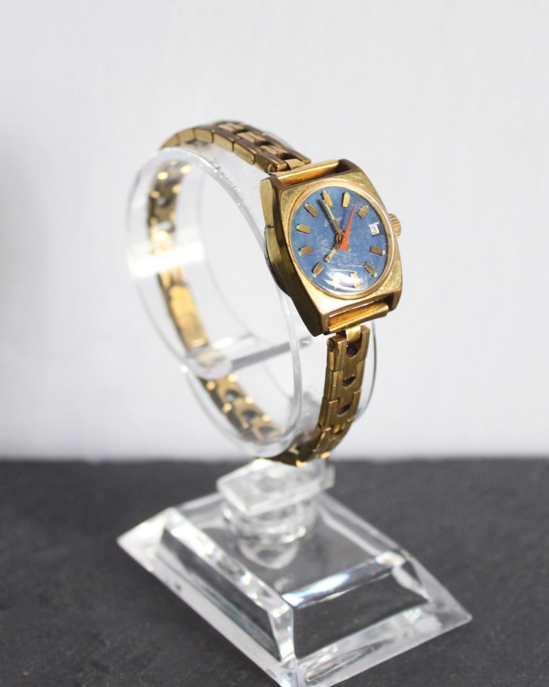 Side view of a functional vintage 1970 gold Latendresse womens mechanical wind up wristwatch. This Stylish watch has a blue dial with golden markers and a contrasting orange hand. It is on a clear bracelet display on white and grey background