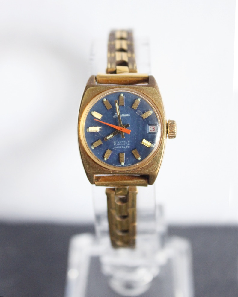 Front view of a functional vintage 1970 gold Latendresse womens mechanical wind up wristwatch. This Stylish watch has a blue dial with golden markers and a contrasting orange hand. It is on a clear bracelet display on white and grey background