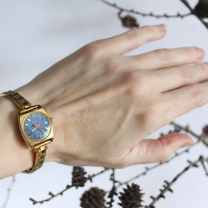 Women left arm with vintage 1970 fully working golden Latendresse mechanical wind up wristwatch. This stylish watch has a blue round dial with gold markers and hands with an orange one. There are branches with small pinecones in the background.