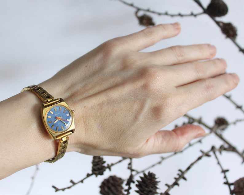 Women left arm with vintage 1970 fully working golden Latendresse mechanical wind up wristwatch. This stylish watch has a blue round dial with gold markers and hands with an orange one. There are branches with small pinecones in the background.