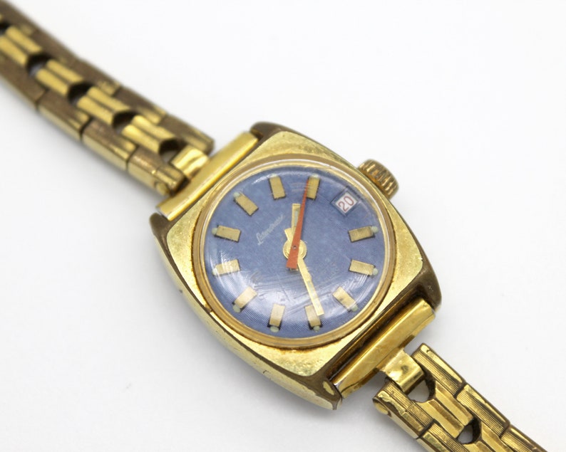 Close up of the dial of a vintage 1970 gold tone Latendresse womens wind up watch. This stylish watch has a blue dial with golden markers and a contrasting vibrant orange hand. It also showcase a date display. The watch informations are written white