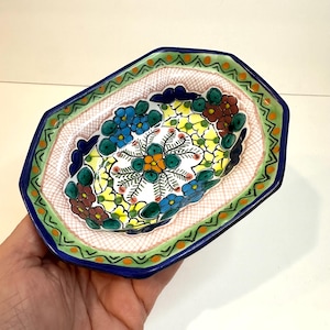 May include: A hand-painted, octagonal ceramic dish with a vibrant floral design. The dish features a blue rim, a green and orange border, and a pink interior with a white floral pattern. The flowers are blue, yellow, and brown.