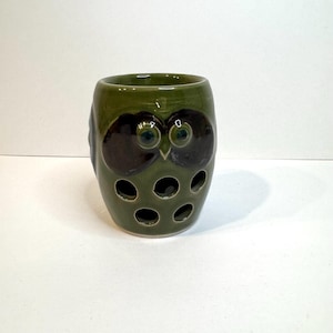 May include: A cylindrical ceramic holder in a glossy olive green finish, shaped like an owl. The front features two large dark brown eye patches with green centers, a small beak, and five circular cutouts arranged in a pattern near the base.