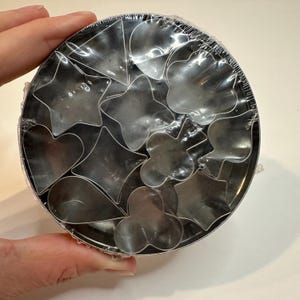 May include: A set of various shaped cookie cutters, including stars, hearts, and clovers, neatly arranged in a round tin. The cutters are made of shiny metal and are packaged in clear plastic wrap.