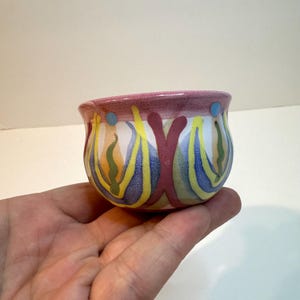 May include: A small ceramic bowl featuring a hand-painted pattern of vertical yellow, blue, and green stripes against a white background, topped with a wide, textured mauve rim accented by small blue dots.