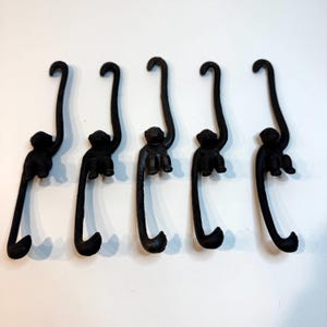 May include: Five black metal monkey-shaped hooks arranged in a row. Each hook features a monkey figure with a long, curved tail that forms a loop at the top and a hook at the bottom, designed for hanging items from the monkey's extended tail.