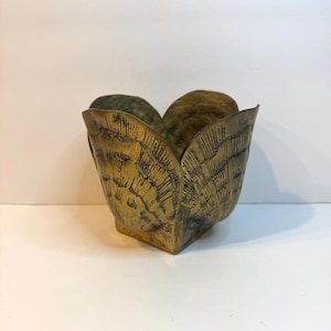 May include: A brass vessel shaped like an open flower or shell, featuring a textured, dark-patina finish with etched lines radiating from the base to the scalloped edges, creating a layered, organic appearance with a metallic gold tone.