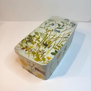 May include: A rectangular metal storage tin featuring a light cream background decorated with a watercolor-style botanical print of green stems, thin brown grasses, small blue flowers, and vibrant yellow blossoms across the lid and sides.