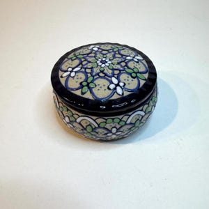 May include: A round ceramic lidded jar featuring a beige base with a hand-painted pattern in dark blue, light green, and white. The lid has a central floral motif surrounded by geometric shapes, while the base displays a repeating scalloped design.