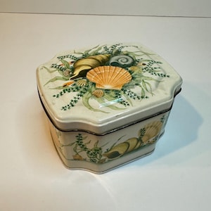 May include: A decorative, hexagonal tin box with a cream-colored background. The lid and sides feature illustrations of seashells, seaweed, and other marine life in shades of yellow, orange, and green. The box has a black trim.