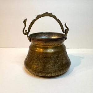 Può includere: Un vaso in ottone con un corpo largo e arrotondato e un collo affusolato. L'esterno presenta intricati motivi incisi e una superficie strutturata. Include un manico in metallo curvo e decorativo fissato a due supporti laterali verticali a forma di serpente.