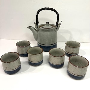 May include: A ceramic tea set featuring a teapot and six matching cups. The teapot has a gray speckled finish with a dark blue band and a woven handle. The cups have a similar design with a blue band and brown rim.