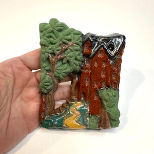May include: A rectangular ceramic wall hanging featuring a detailed relief of a landscape. The scene includes a red building with a black roof, trees, and a winding path. The colors are primarily green, brown, and red.