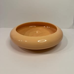 May include: A round, shallow ceramic bowl with a smooth, glossy finish in a solid light peach color. The bowl features a wide, rounded rim that curves inward toward the center, creating a simple, circular basin with a uniform, reflective surface.
