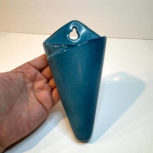 May include: A cone-shaped wall planter with a smooth, teal-colored glazed finish. The ceramic vessel features a pointed base and a flat top section with a keyhole-shaped opening designed for mounting on a wall surface for display purposes.