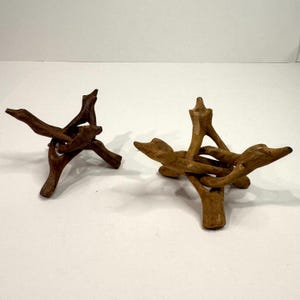 May include: Two tripod display stands crafted from interlocking pieces of natural brown wood. Each stand features three legs and three upward-pointing arms designed to support spherical objects, showcasing a raw, organic texture and finish.