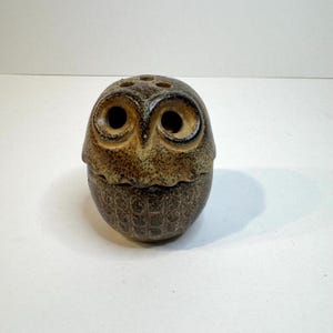 May include: A brown ceramic owl figurine with a speckled glaze finish. The owl features large, circular eye sockets, a pointed beak, and a textured body with a repeating scalloped pattern. Three small circular holes are visible on the top of the head.