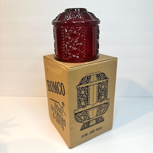 Pode incluir: Um frasco de vidro vermelho rubi com tampa, com um design floral em relevo. O frasco está em cima de uma caixa de cartão com o logotipo HOMCO e o texto "HOME INTERIORS & GIFTS". A caixa também exibe uma ilustração de um frasco semelhante.