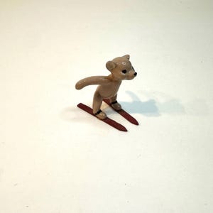 May include: A small, tan-colored teddy bear figurine skiing downhill on red skis. The bear has black eyes and is posed with arms outstretched. The skis are a dark red color.