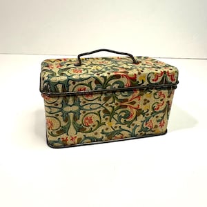 Puede incluir: Una caja rectangular de metal para guardar cosas con un estampado floral en rojo, verde y amarillo sobre un fondo crema. Tiene un asa y ribetes de metal oscuro. El recipiente vintage es adecuado para guardar objetos pequeños. Mide unos 15 cm de largo.