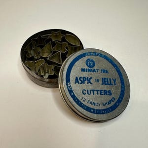 May include: An open, round, metal tin containing various miniature cookie cutters in star, heart, and other shapes. The lid of the tin is visible, with blue text that reads "Miniature Aspic or Jelly Cutters, 12 Fancy Shapes."