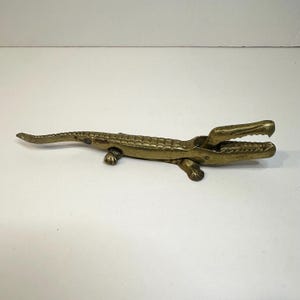 Może przedstawiać: Mosiężny dziadek do orzechów w kształcie aligatora z otwartą paszczą, szczegółowymi łuskami i czterema małymi nóżkami. Dekoracyjne narzędzie kuchenne.