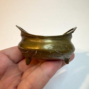May include: A small, antique-style brass bowl featuring a rounded body, three short legs, and two upward-curving handles on opposite sides. The exterior surface of the brass vessel displays etched, linear patterns and a weathered, metallic patina.