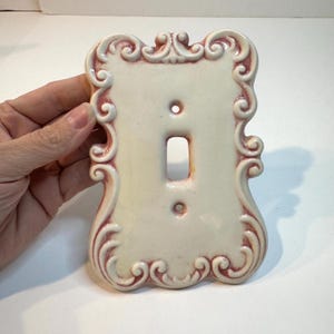 May include: A cream-colored ceramic light switch plate featuring a raised, ornate scrollwork border with pink detailing. The rectangular plate has a central vertical cutout for a toggle switch and two small circular holes for mounting screws.