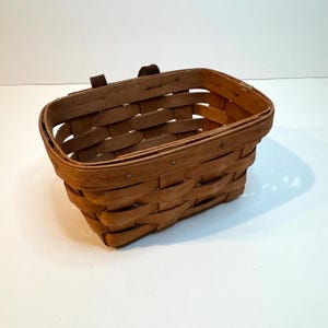 Puede incluir: Una cesta rectangular de madera tejida con un acabado marrón natural. La cesta presenta un borde resistente, una base plana y dos pequeñas trabillas de sujeción de cuero marrón oscuro fijadas en el borde superior con listones entrelazados.