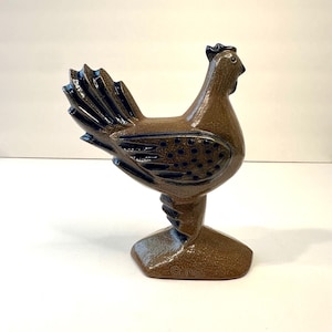 Puede incluir: Figura de gallo de cerámica marrón con detalles en azul oscuro en las plumas de la cola y el cuerpo. El gallo está sobre una base de pedestal. El año "© 1985" está impreso en la base.