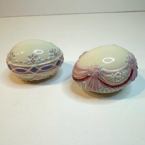 May include: Two decorative egg-shaped trinket boxes. One is cream-colored with purple and blue accents, the other has pink and gold trim with floral details. Both have a glossy finish.