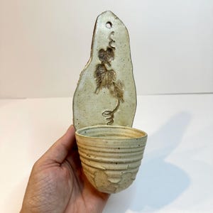 May include: Handmade ceramic wall pocket planter with a natural, earthy aesthetic. The planter features a textured, light-colored background with a decorative embossed vine design. The attached cup-shaped holder is designed to hold small plants or flowers.